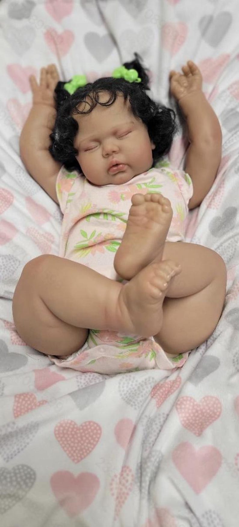 May include: A lifelike baby doll with dark hair and green bows, dressed in a pink floral onesie, rests on a blanket patterned with pink and grey hearts. The doll's arms are raised, and its eyes are closed.