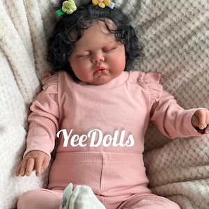 May include: A lifelike baby doll wearing a pink ribbed long-sleeved top and matching pants. The doll has dark curly hair, closed eyes, and is adorned with flower hair clips. The text "YeeDolls" is printed on the clothing.