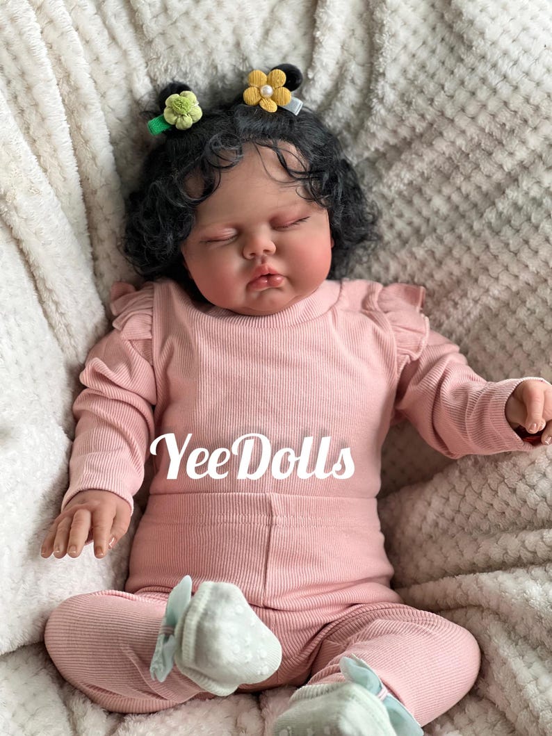 May include: A lifelike baby doll wearing a pink ribbed long-sleeved top and matching pants. The doll has dark curly hair, closed eyes, and is adorned with flower hair clips. The text "YeeDolls" is printed on the clothing.