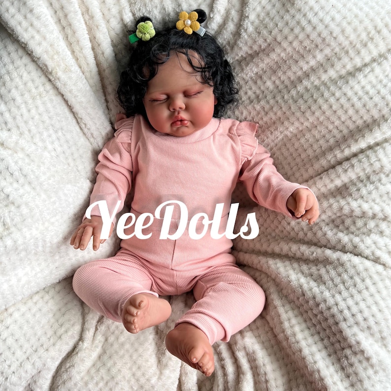 May include: A lifelike baby doll with dark curly hair, wearing a pink ribbed long-sleeved top and matching pants. The doll has closed eyes and is adorned with small flower hair clips. The text "YeeDolls" is displayed across the doll.