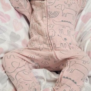 May include: A realistic baby doll in a pink onesie with a grey animal print, resting on a soft blanket. The doll has dark hair and a pink bow. The blanket features a heart pattern.