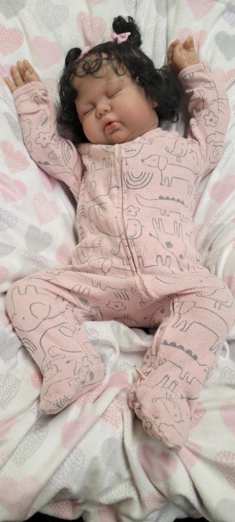 May include: A realistic baby doll in a pink onesie with a grey animal print, resting on a soft blanket. The doll has dark hair and a pink bow. The blanket features a heart pattern.