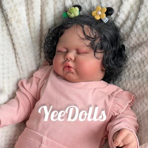 May include: A lifelike baby doll with dark curly hair and closed eyes, wearing a pink long-sleeved shirt. The doll has two small flower hair clips. The text "Yee Dolls" is printed on the shirt.