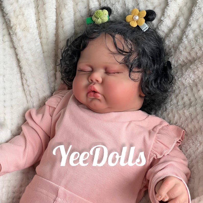 May include: A lifelike baby doll with dark curly hair and closed eyes, wearing a pink long-sleeved shirt. The doll has two small flower hair clips. The text "Yee Dolls" is printed on the shirt.
