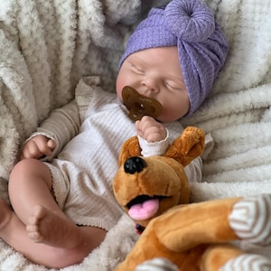 May include: A sleeping baby doll wearing a lavender turban and a pacifier. The doll is dressed in a cream-colored onesie and is resting on a fluffy white blanket. A brown stuffed dog toy is next to the doll.