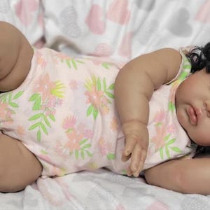 May include: A realistic baby doll with dark hair in pigtails, adorned with green bows. The doll is dressed in a light pink onesie featuring a floral design and rests on a patterned blanket. The doll's eyes are closed, suggesting sleep.