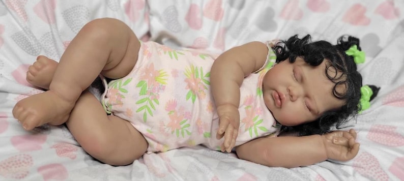 May include: A realistic baby doll with dark hair in pigtails, adorned with green bows. The doll is dressed in a light pink onesie featuring a floral design and rests on a patterned blanket. The doll's eyes are closed, suggesting sleep.