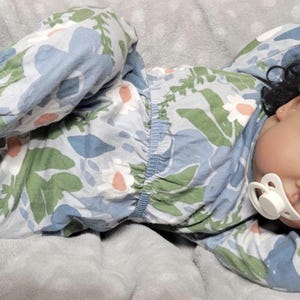 May include: A realistic baby doll dressed in a floral patterned pyjama set in blue, green, and white, with a dummy in its mouth. The doll has dark curly hair and red bows. The doll is lying on a grey blanket.