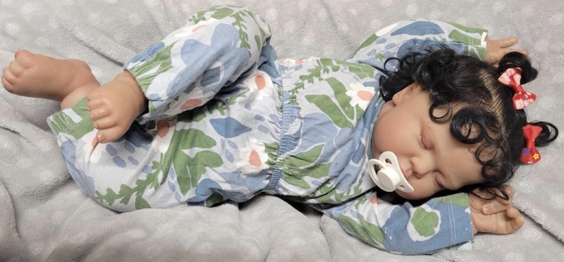 May include: A realistic baby doll dressed in a floral patterned pyjama set in blue, green, and white, with a dummy in its mouth. The doll has dark curly hair and red bows. The doll is lying on a grey blanket.