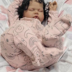 May include: A realistic baby doll with dark curly hair and closed eyes, dressed in a pink onesie featuring a grey animal and rainbow design. The doll rests on a patterned blanket with pink and grey hearts.