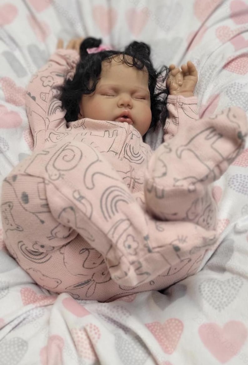 May include: A realistic baby doll with dark curly hair and closed eyes, dressed in a pink onesie featuring a grey animal and rainbow design. The doll rests on a patterned blanket with pink and grey hearts.