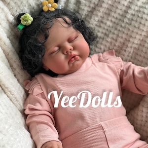 May include: A lifelike baby doll with dark curly hair, wearing a pink ribbed outfit. The doll has closed eyes and is adorned with flower hair clips. The text "YeeDolls" is displayed on the outfit.