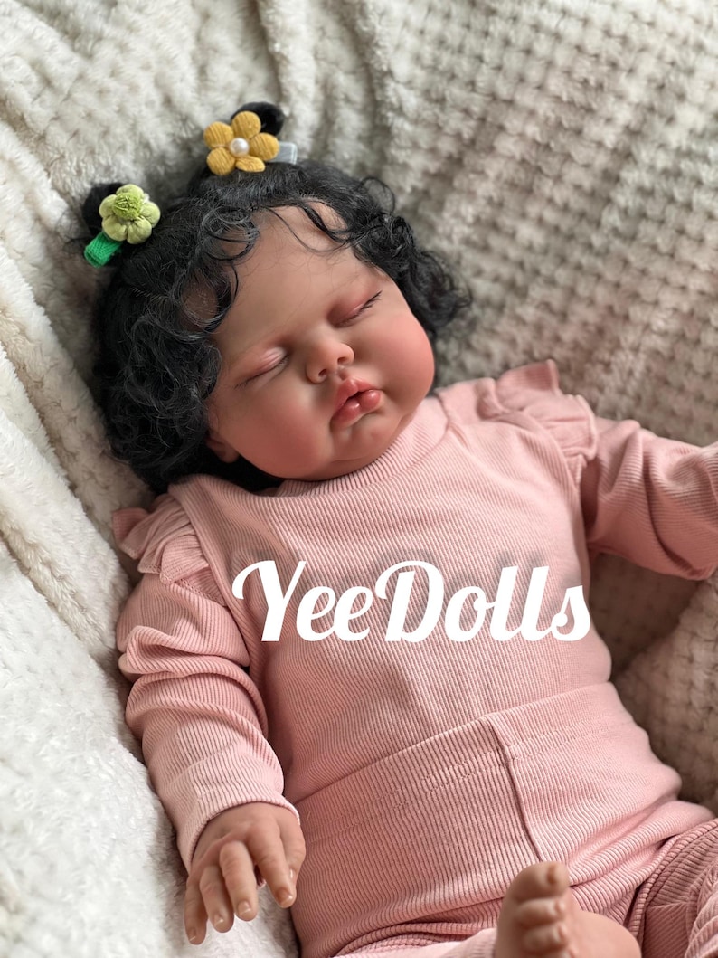 May include: A lifelike baby doll with dark curly hair, wearing a pink ribbed outfit. The doll has closed eyes and is adorned with flower hair clips. The text "YeeDolls" is displayed on the outfit.