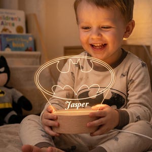 May include: A custom acrylic night light featuring an illuminated Batman logo outline and the name Jasper engraved on the front. The clear acrylic piece is set into a round, light-coloured wooden base with a power cord extending from the side.