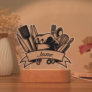 May include: A wooden-based night light with a black and white chef's hat and kitchen utensil design. The name "Jane" is displayed on a banner. The light is on, illuminating the design.