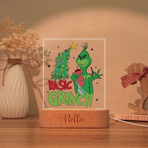 May include: A decorative night light featuring a green Grinch character holding a red drink, with a Christmas tree and the words "BASIC GRINCH". The light sits on a wooden base with the name "Nelle" engraved.