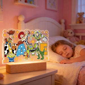 Peut inclure: Une veilleuse en acrylique éclairée avec des personnages du film Toy Story. La lumière est montée sur une base en bois et épelle le mot "NAME" avec les images des personnages. La lumière est sur une table de chevet à côté d'un enfant endormi.