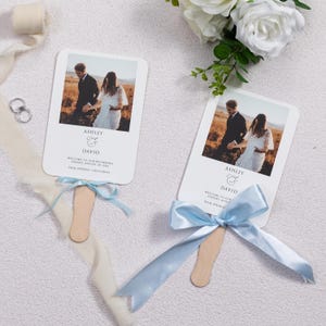 Peut inclure: Deux éventails de mariage blancs avec une photo d'un couple, les noms "Ashley & David" et les détails du mariage. Chaque éventail a un ruban bleu clair et une poignée en bois. Les éventails sont sur une surface blanche avec des fleurs et des rubans.