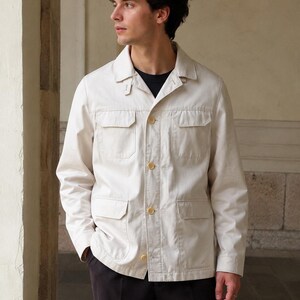 Può includere: Una giacca utility color beige chiaro con colletto a punta, maniche lunghe e quattro tasche frontali con patta. La giacca si chiude con quattro bottoni rotondi di colore chiaro sul davanti e presenta un piccolo dettaglio a linguetta sul colletto.