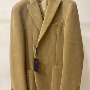 Op de afbeelding: Een beige corduroy blazer met een reverskraag, twee voorzakken en drie knopen op elke manchet. De jas hangt aan een zwarte hanger. Er is een label aan de jas bevestigd.