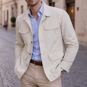 Puede incluir: Chaqueta beige claro con cuatro bolsillos delanteros, usada sobre una camisa azul claro. El atuendo incluye pantalones color caqui y un cinturón de cuero marrón. Se ve un reloj plateado en la muñeca.