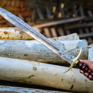 May include: A silver sword with a damascus steel blade and a brown wooden handle. The sword is resting on a pile of logs.