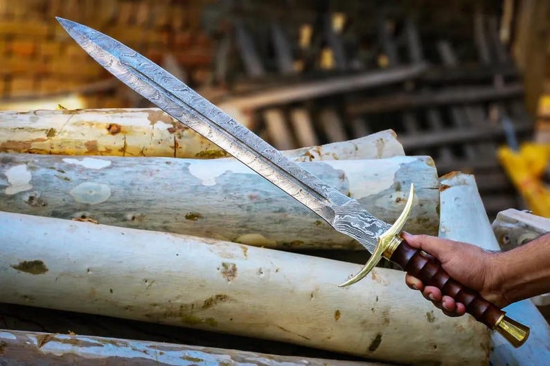 May include: A silver sword with a damascus steel blade and a brown wooden handle. The sword is resting on a pile of logs.