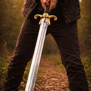 May include: A long, silver sword with a gold hilt is held upright. The sword's blade is polished, reflecting light. The hilt features ornate gold detailing. The sword is held in front of a person, with a blurred forest background.