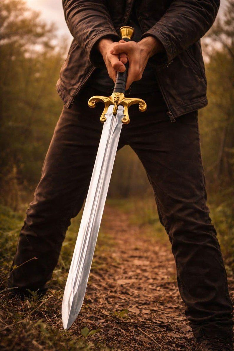 May include: A long, silver sword with a gold hilt is held upright. The sword's blade is polished, reflecting light. The hilt features ornate gold detailing. The sword is held in front of a person, with a blurred forest background.