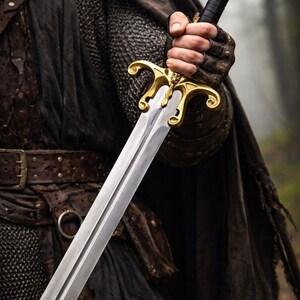 May include: A silver sword with a gold hilt and black handle is held in a gloved hand. The sword has a double-edged blade and ornate gold detailing. The person is wearing a dark cloak and leather armor.