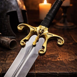 May include: A detailed image of a decorative sword with a gold-colored hilt and a skull design. The blade is silver, and the handle is wrapped in black leather. The sword rests on a wooden surface, with candles and a helmet in the background.