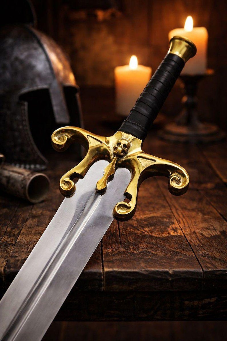 May include: A detailed image of a decorative sword with a gold-colored hilt and a skull design. The blade is silver, and the handle is wrapped in black leather. The sword rests on a wooden surface, with candles and a helmet in the background.