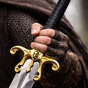 May include: A close-up of a sword with a gold-colored hilt featuring a skull design. The handle is wrapped in black leather. The blade is silver and reflects light. The sword is held by a person wearing brown leather gloves.