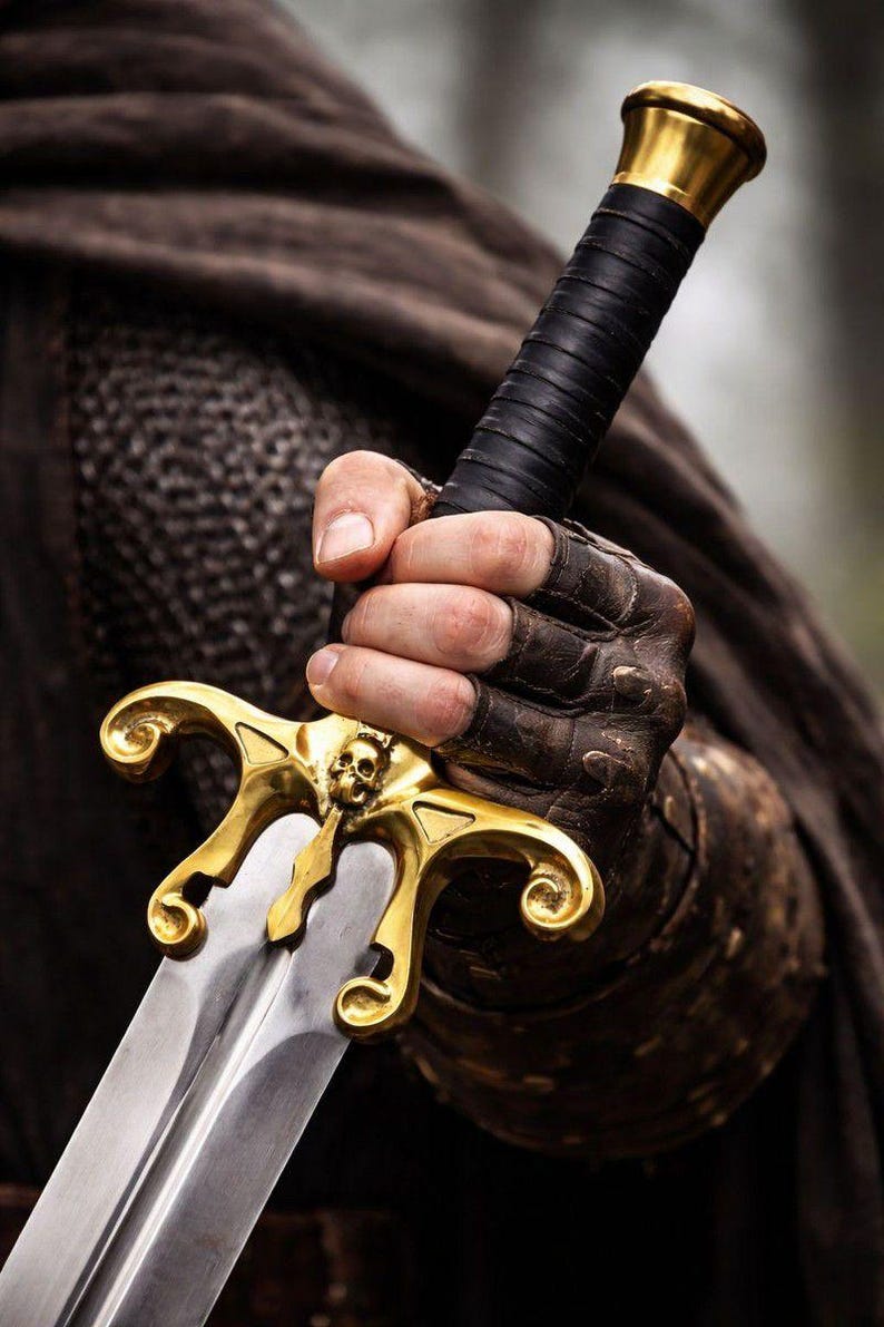 May include: A close-up of a sword with a gold-colored hilt featuring a skull design. The handle is wrapped in black leather. The blade is silver and reflects light. The sword is held by a person wearing brown leather gloves.