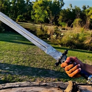 May include: A silver sword with a patterned blade and a black handle with gold accents. The sword is held by a hand in a grassy field.