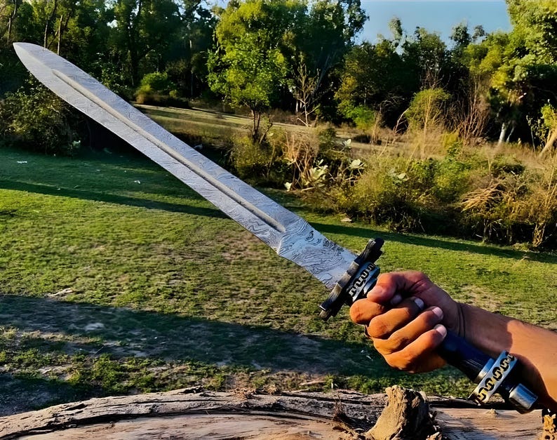 May include: A silver sword with a patterned blade and a black handle with gold accents. The sword is held by a hand in a grassy field.