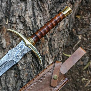 May include: A sword with a brown wooden handle and a silver blade with a decorative pattern. The sword is resting on a brown leather sheath with a decorative pattern.