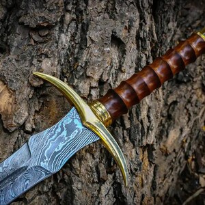 May include: A sword with a blue and silver patterned blade, a brown wooden handle, and a gold-colored pommel and guard. The sword is resting against a tree trunk.