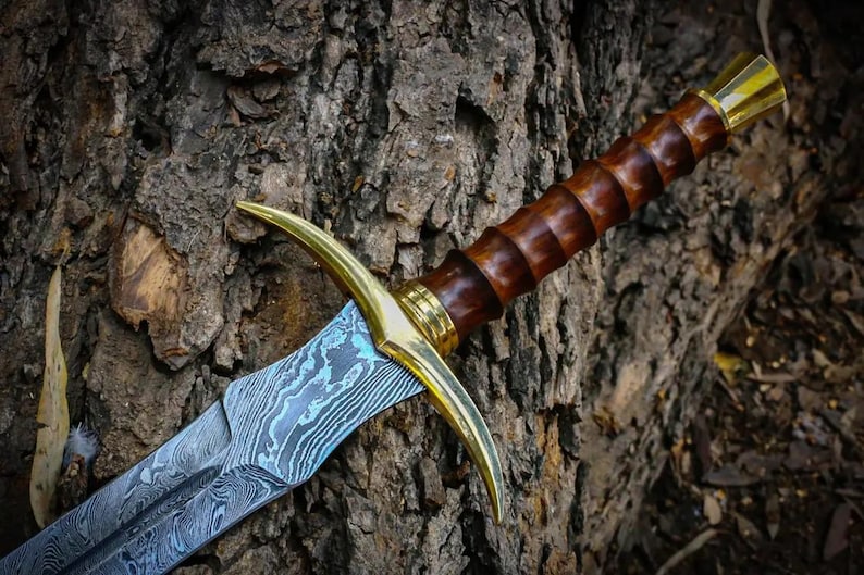 May include: A sword with a blue and silver patterned blade, a brown wooden handle, and a gold-colored pommel and guard. The sword is resting against a tree trunk.