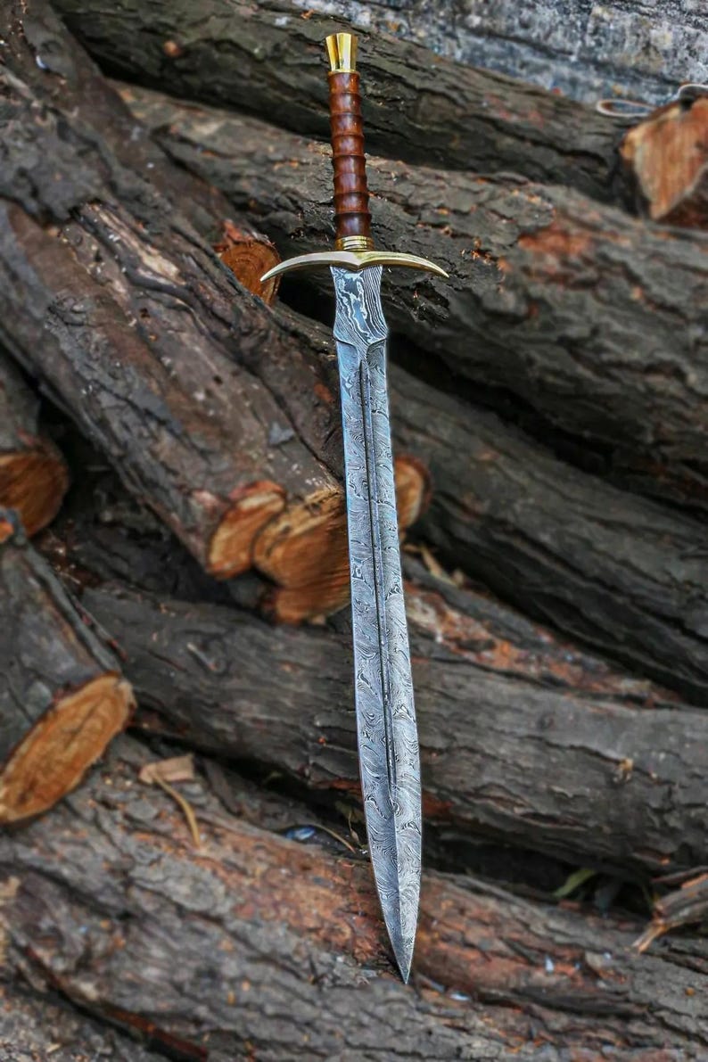 May include: A long, silver sword with a wooden handle and a gold pommel. The sword is lying on a pile of logs.