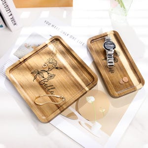 Op de afbeelding: Twee houten trays met gegraveerde bloemmotieven en namen. E&eacute;n tray bevat een gouden ketting en armband, de andere een zilveren horloge en munten. De trays liggen op een wit oppervlak met de tekst "You have only one life."