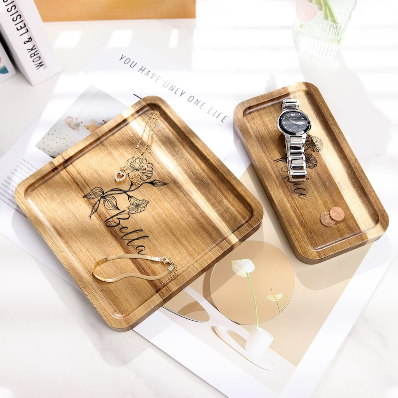 Op de afbeelding: Twee houten trays met gegraveerde bloemmotieven en namen. E&eacute;n tray bevat een gouden ketting en armband, de andere een zilveren horloge en munten. De trays liggen op een wit oppervlak met de tekst "You have only one life."