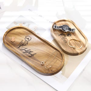 Op de afbeelding: Twee ovale houten trays met gegraveerde bloemmotieven en namen. E&eacute;n tray bevat een horloge en een armband, de andere een ketting en een munt. De trays zijn gemaakt van lichtbruin hout.
