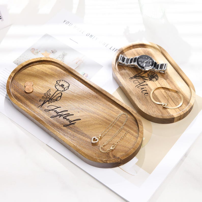 Op de afbeelding: Twee ovale houten trays met gegraveerde bloemmotieven en namen. E&eacute;n tray bevat een horloge en een armband, de andere een ketting en een munt. De trays zijn gemaakt van lichtbruin hout.