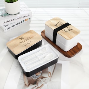 May include: Two black and white lunch boxes with wooden lids. The lids have the text "Olivia's Lunch" and "Sean's Lunch" engraved on them. The lunch boxes come with a set of white utensils.