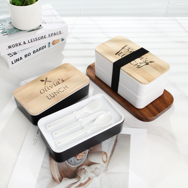 May include: Two black and white lunch boxes with wooden lids. The lids have the text "Olivia's Lunch" and "Sean's Lunch" engraved on them. The lunch boxes come with a set of white utensils.