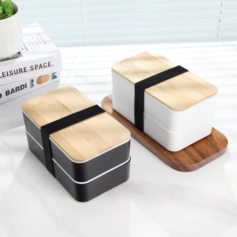 May include: Two black and white lunch boxes with wooden lids and black elastic bands. The lunch boxes are on a wooden surface.