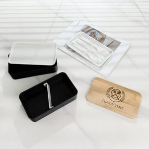 May include: A black lunchbox with a white lid and a separate compartment for utensils. The lunchbox has a wooden lid with the text "Lunch Time" and a graphic of a fork and knife.