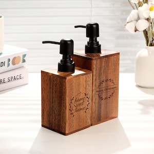 Può includere: Due dispenser di sapone in legno con pompe nere. Uno ha le parole "home sweet home" in un carattere decorativo. L'altro ha le parole "Merry Christmas" in un carattere decorativo. I dispenser sono in legno scuro.