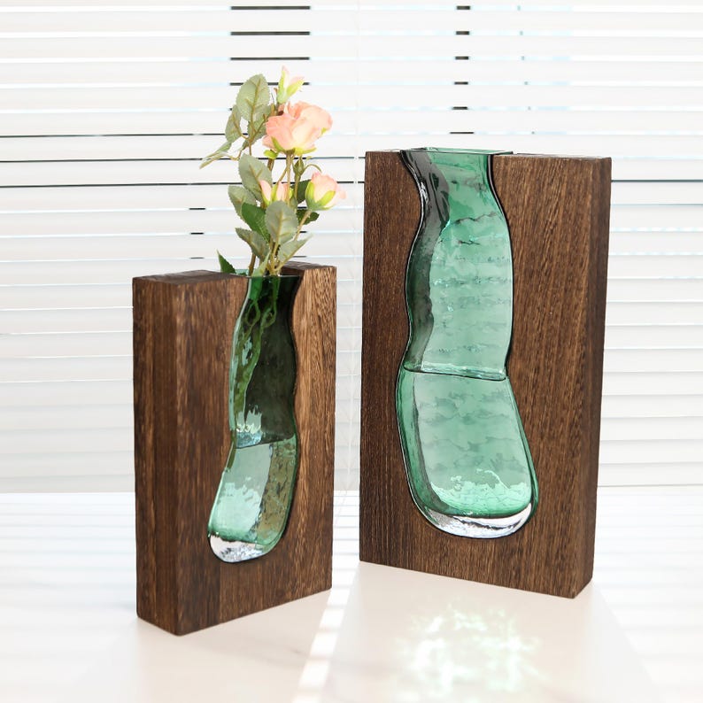 May include: Two wooden vases with a dark brown finish. Each vase has a unique, curved, teal-coloured glass insert. One vase holds pink flowers with green leaves. The vases are displayed against a white background.